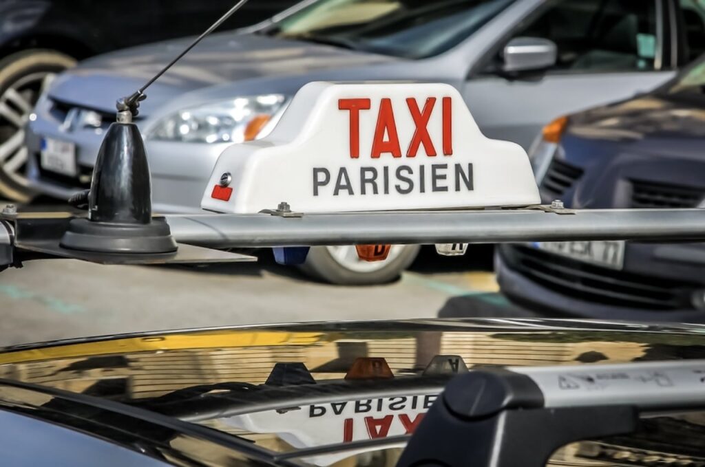 Close-up of a white illuminated 'TAXI PARISIEN' sign on the roof of a taxi with a blurred background of other vehicles, depicting a busy urban environment in Paris, France.