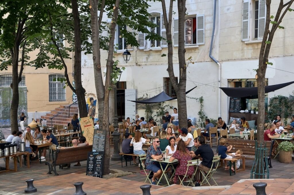 Vibrant outdoor cafe scene in Marseille, France, with people enjoying drinks and conversation at wooden tables amidst lush trees and historic architecture, conveying the relaxed atmosphere of European street dining.