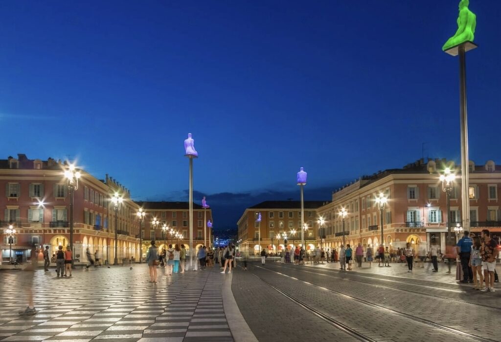 Twilight descends on Place Masséna in Nice, where locals and tourists alike stroll among the checkerboard pavement and illuminated statue-lined square, a must-visit landmark in a 'Nice in one day' tour.