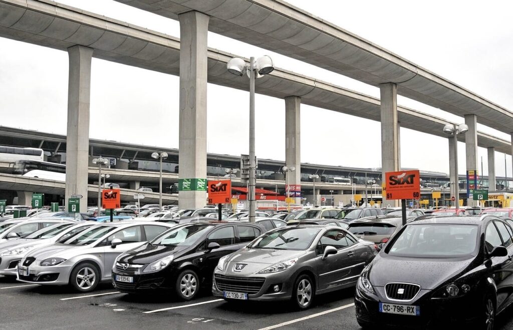 A packed rental car parking lot at an airport with multiple signs for various rental companies such as Sixt, Avis, and Hertz, with an overpass and part of an airport terminal in the background.