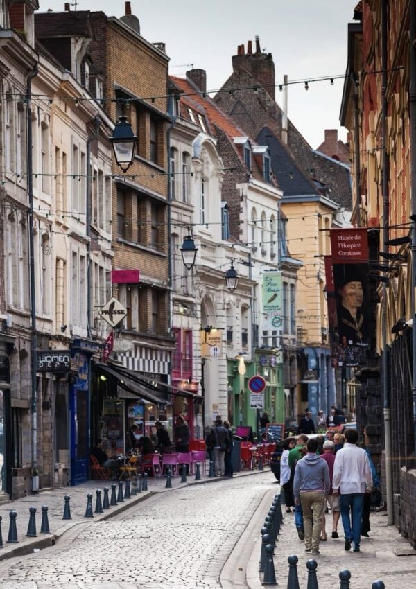 Cobbled street scene in Lille with pedestrians walking past traditional European buildings, vibrant shopfronts, and street lamps, leading towards the Musée de l'Hospice Comtesse sign in the background.