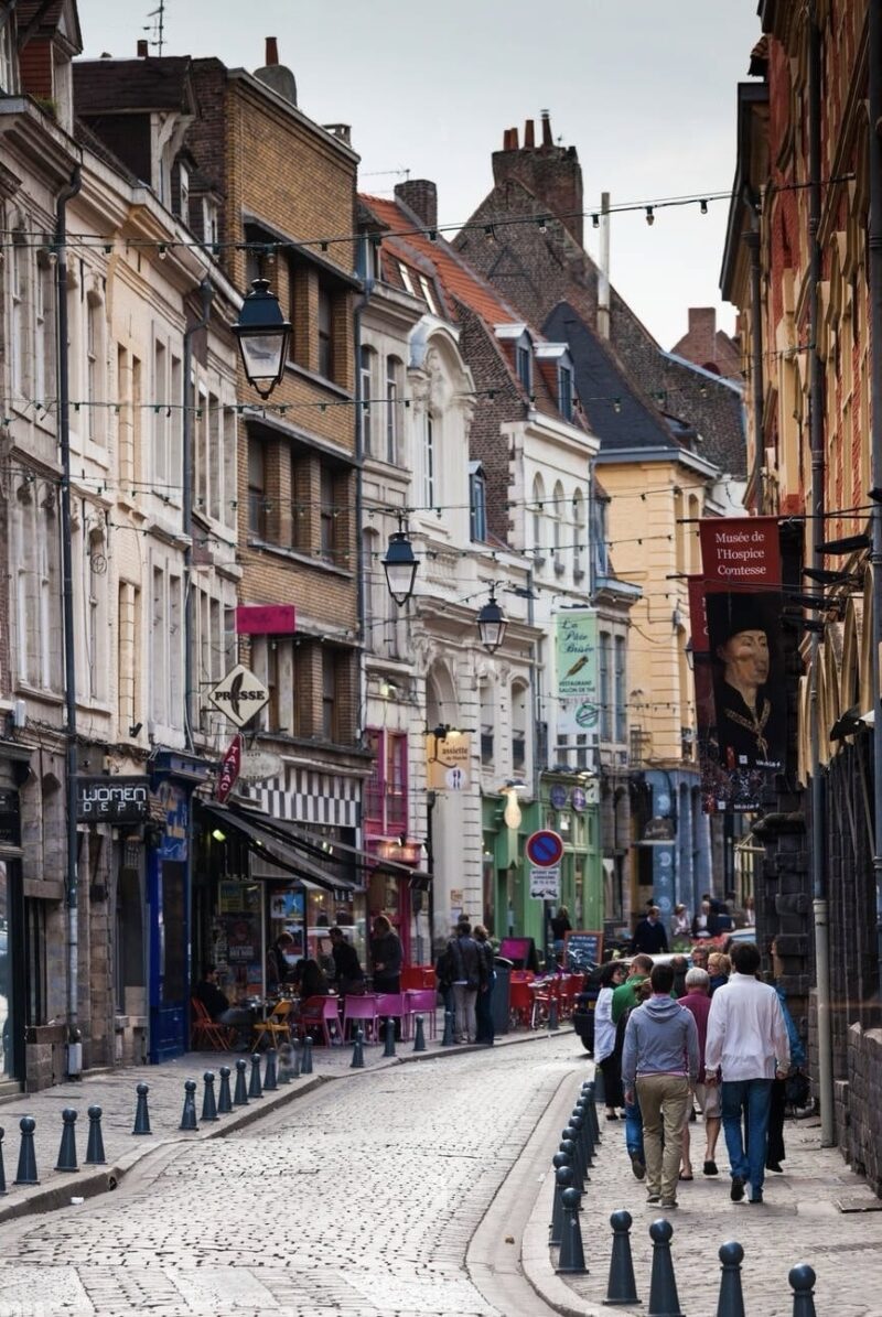 Cobbled street scene in Lille with pedestrians walking past traditional European buildings, vibrant shopfronts, and street lamps, leading towards the Musée de l'Hospice Comtesse sign in the background.