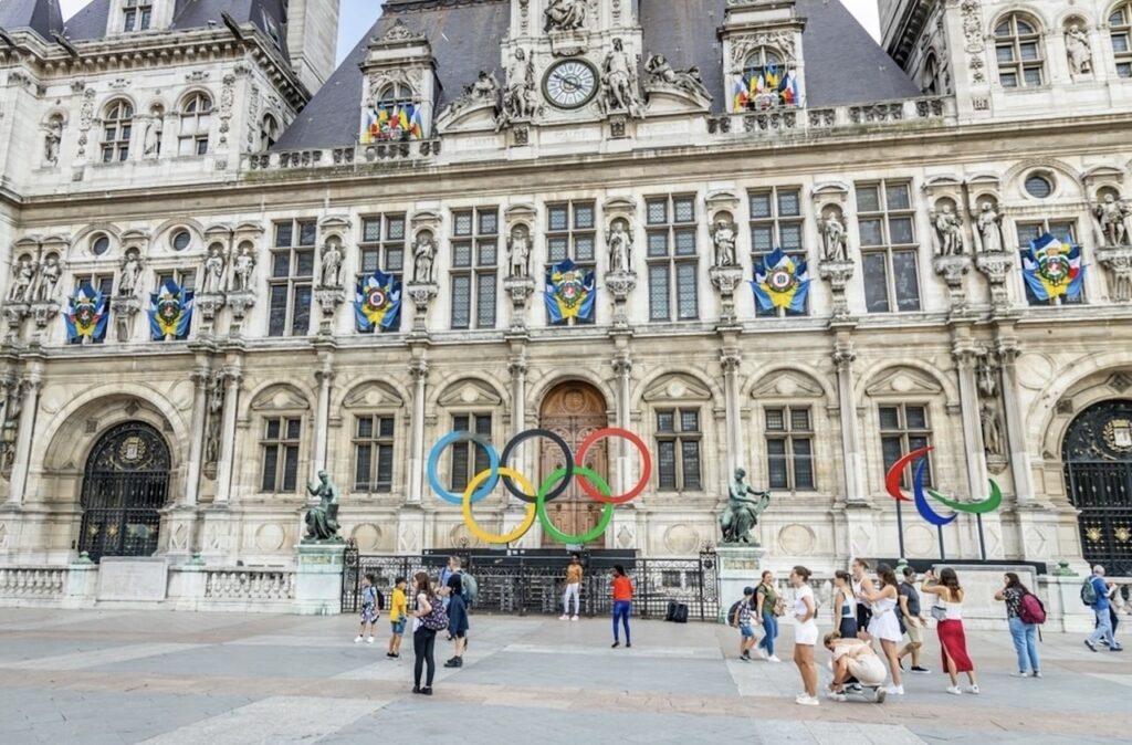 Tourists gather in the square of Hôtel de Ville in Paris, with the iconic Olympic rings installation in vibrant colors, celebrating the forthcoming games, set against the ornate façade of the city hall adorned with flags.