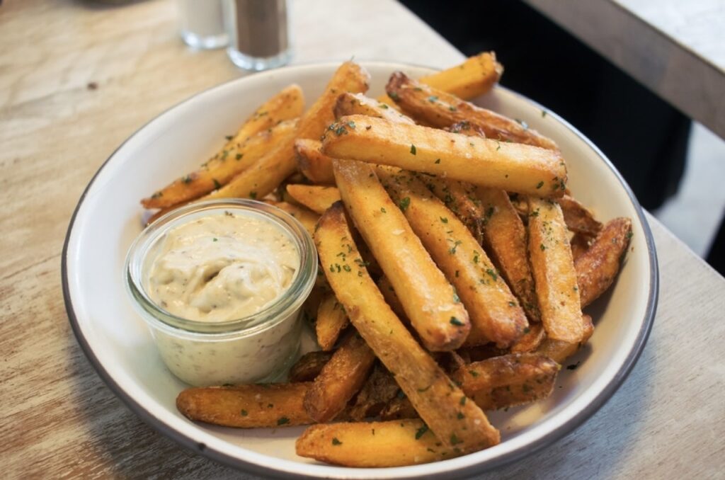 A bowl of golden truffle fries sprinkled with parsley, served with a side of aioli sauce, placed on a wooden table, capturing the essence of a casual, yet indulgent, snack experience.