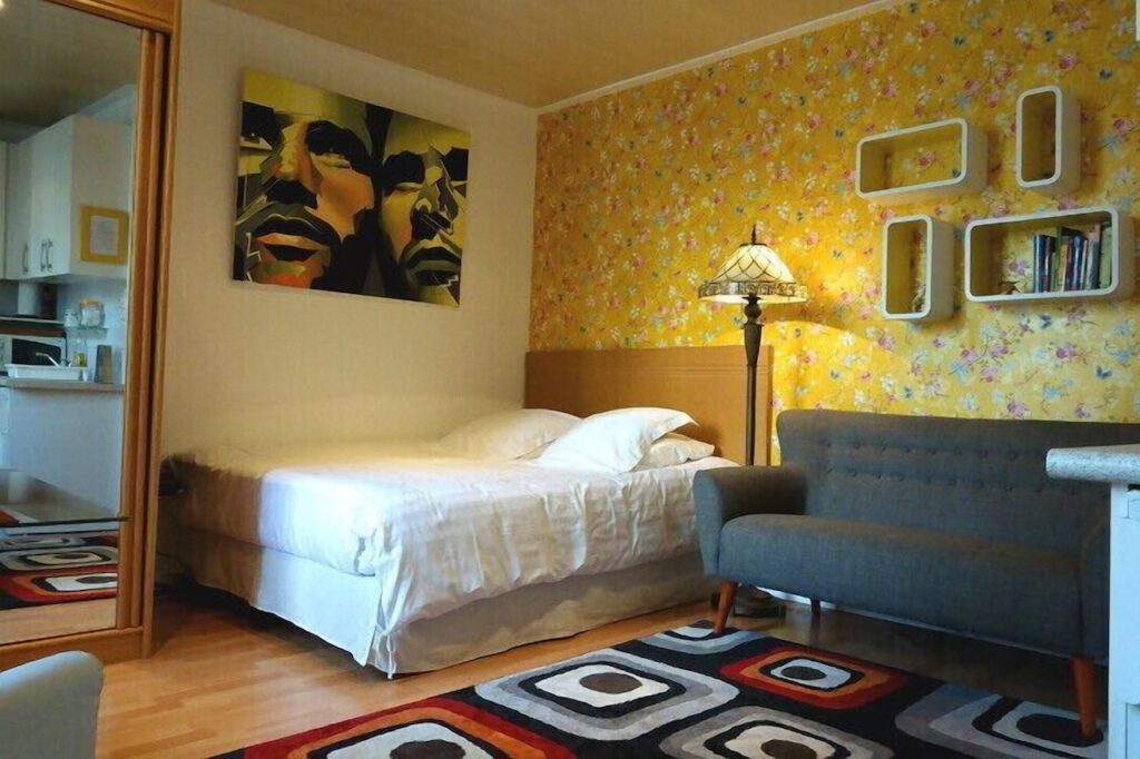 Warm and inviting bedroom with a vibrant yellow floral wallpaper, a modern abstract painting above the bed, a classic floor lamp, and a geometric patterned rug complementing a gray sofa.