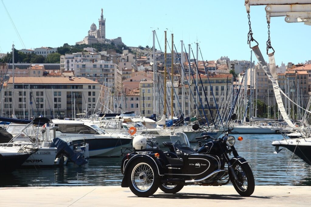 A classic black motorcycle with a sidecar parked at a marina in Marseille, France, with an array of sailboats docked in the background and the historic Notre-Dame de la Garde basilica perched on a hill, highlighting a scenic spot for motorcycle tours in France.