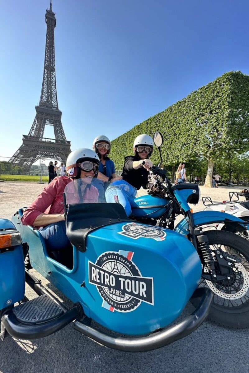 Three happy tourists wearing helmets are seated on blue sidecar motorcycles with the 'Retro Tour Paris' logo, enjoying a sunny day with the iconic Eiffel Tower in the background, on their motorcycle tour in France.