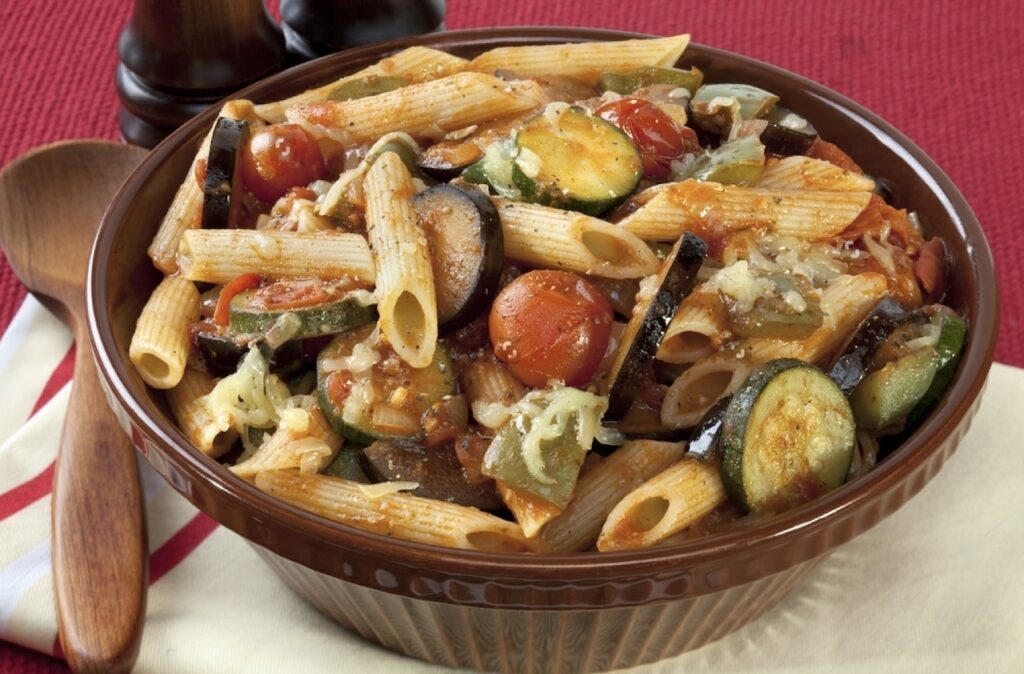 A hearty bowl of penne pasta mixed with zucchini, eggplant, cherry tomatoes, and garnished with shredded cheese, served on a wooden table with a red napkin."This description conveys the contents of the bowl, the type of pasta, the main vegetables included, and the garnishing, while also providing context about the setting.