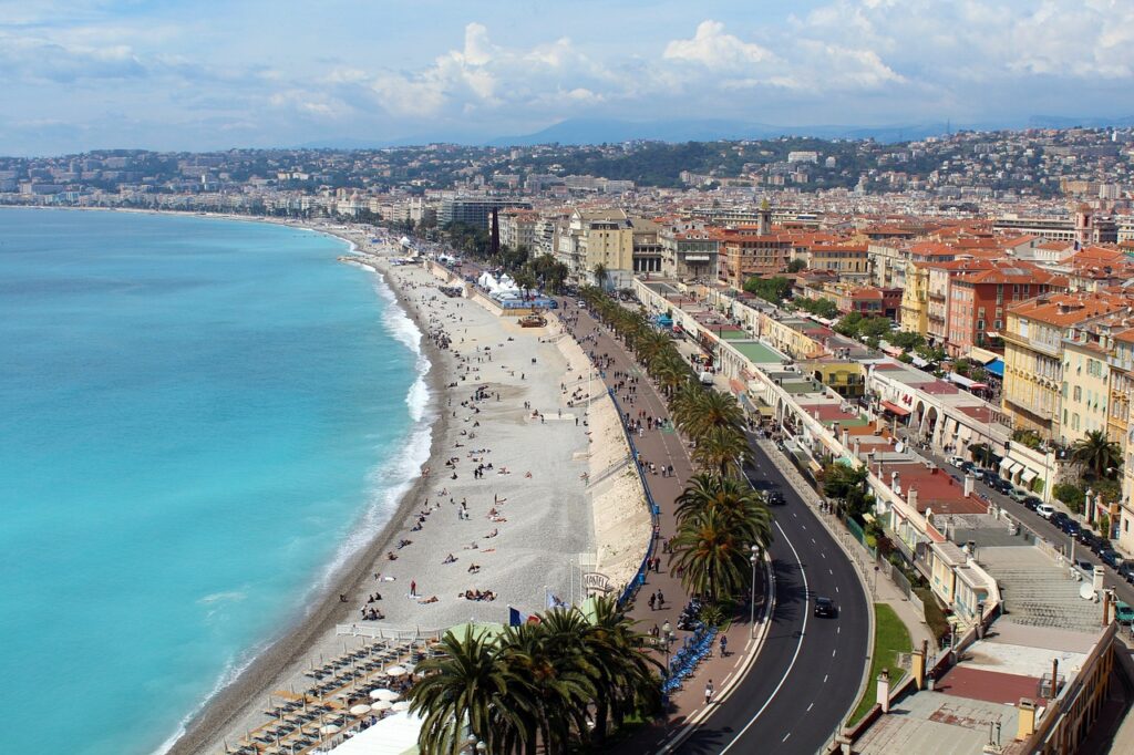 Aerial view of Nice's Promenade des Anglais, with its iconic pebble beach along the azure Mediterranean Sea, lined with palm trees and Belle Époque architecture, a defining image of the best cities in the South of France.