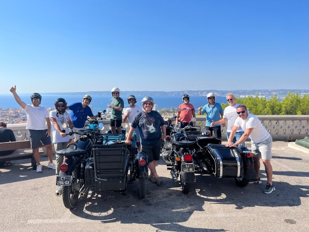 A cheerful group of eight people posing with their black sidecar motorcycles against a panoramic backdrop of the coastline in France, capturing the essence of camaraderie on a motorcycle tour in France.