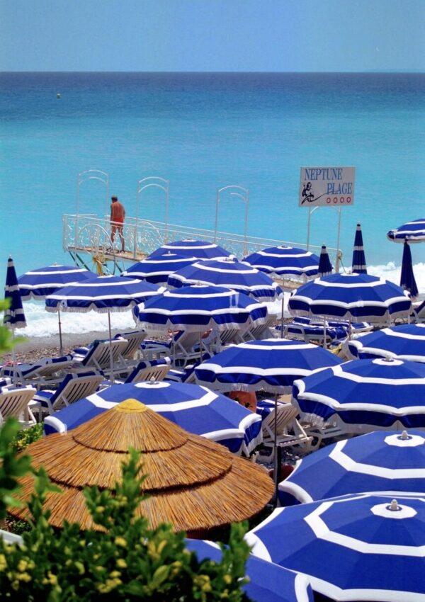 A vibrant seaside atmosphere at one of the beach clubs in Nice, with a row of neatly arranged blue and white umbrellas providing shade to white sun loungers. The brilliant blue ocean stretches out in the background, meeting the clear sky on the horizon. A quaint 'Neptune Plage' sign adds a touch of charm to the scene, while a solitary figure stands on a pier overlooking the Mediterranean Sea, embodying the relaxed and luxurious spirit of Nice's shoreline retreats.