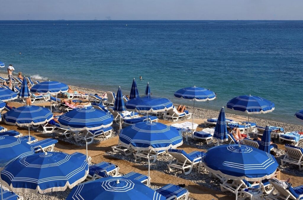 A bustling beach clubs in Nice features rows of striped blue umbrellas and matching loungers on the pebbly shore, with beachgoers enjoying the sun and sea views.