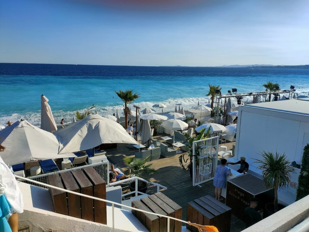 An elegant beach clubs in Nice overlooks the turquoise sea, featuring white umbrellas and loungers. Patrons relax and socialize in this chic, palm-adorned seaside venue, capturing the sophisticated leisure culture of the French Riviera.