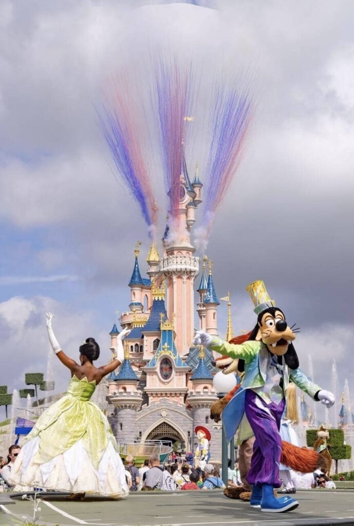 A lively Disneyland Paris parade in front of the majestic Sleeping Beauty Castle, featuring Princess Tiana and Goofy dancing, with a spectacular daytime fireworks display, encapsulating 'Disneyland Paris on a Budget' festivities.