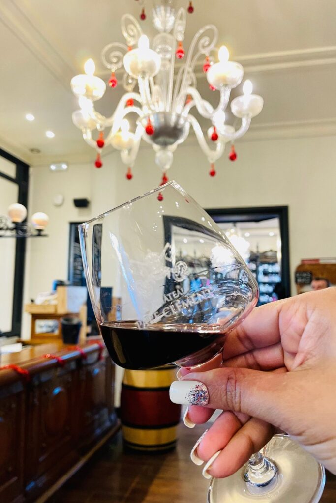 A person's hand holding a glass of rich, red wine from Château Pape Clément, a luxurious touch to any one day in Bordeaux experience. In the softly focused background, the elegant interior of a wine tasting room with a classic white chandelier creates an inviting atmosphere.