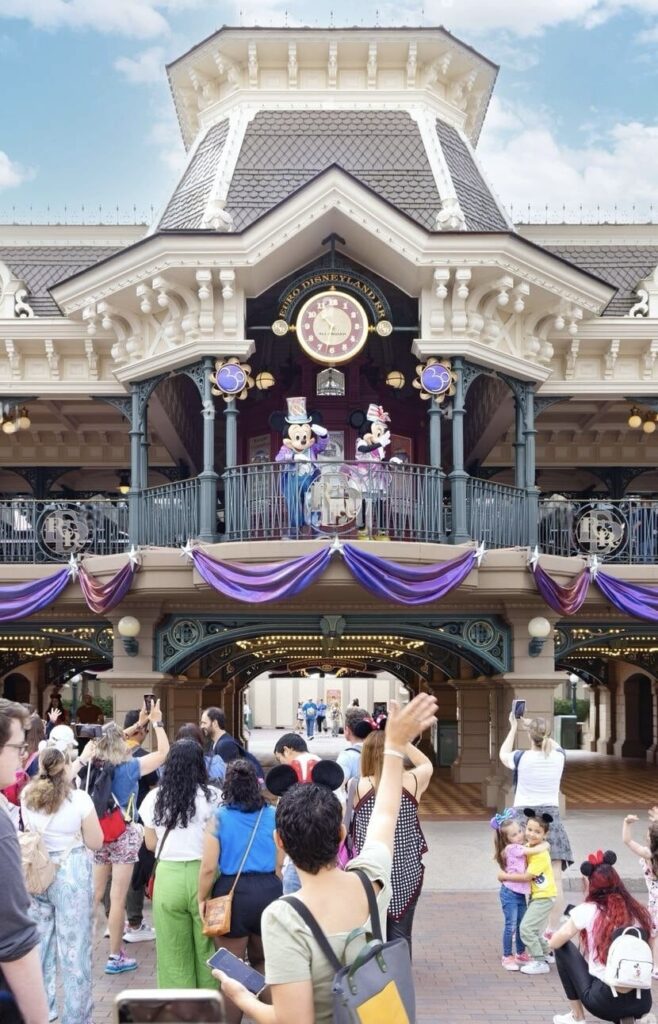 Excited guests waving at Mickey and Minnie Mouse who are standing on the balcony of the Disneyland Paris entrance, adorned with elegant purple drapery, as part of an enchanting 'Disneyland Paris on a Budget' welcome.