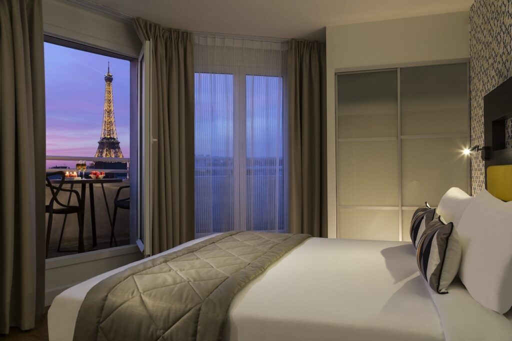 Modern hotel room featuring a large bed with white linens and decorative pillows, a set of curtains framing a window with a breathtaking view of the Eiffel Tower at dusk, emphasizing hotels in Paris with Eiffel Tower views.
