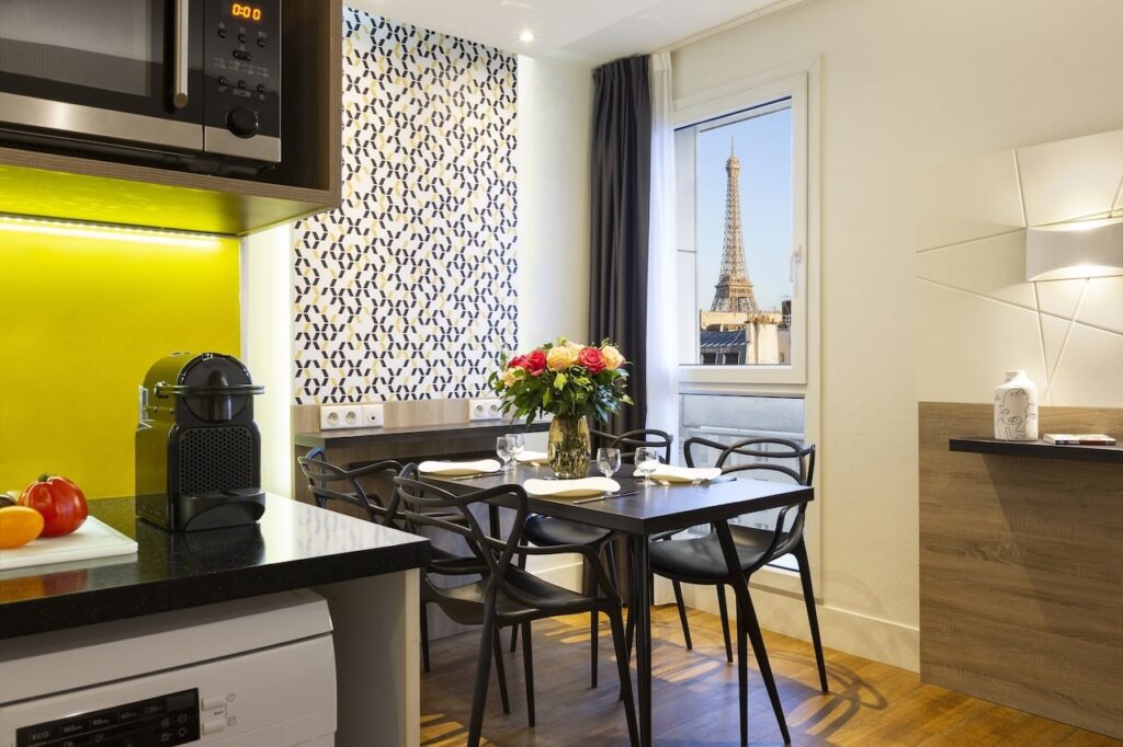 Chic kitchenette with vibrant yellow backsplash in a Paris hotel, featuring a sleek black dining table set with a vase of roses, and a view of the Eiffel Tower from the adjacent window, perfect for guests searching for hotels in Paris with Eiffel Tower views.