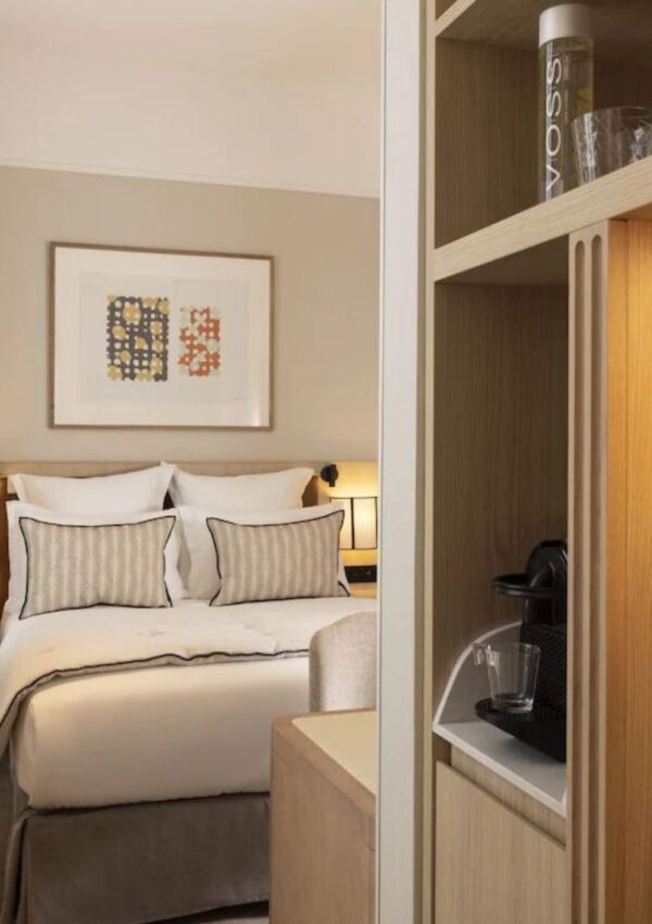 Elegant Paris hotel room featuring a plush double bed with striped pillows, ambient lighting, and artwork on the wall, as viewed from a connecting room that includes a shelf with a VOSS water bottle. Ideal for travelers searching for Paris hotels with connecting rooms.