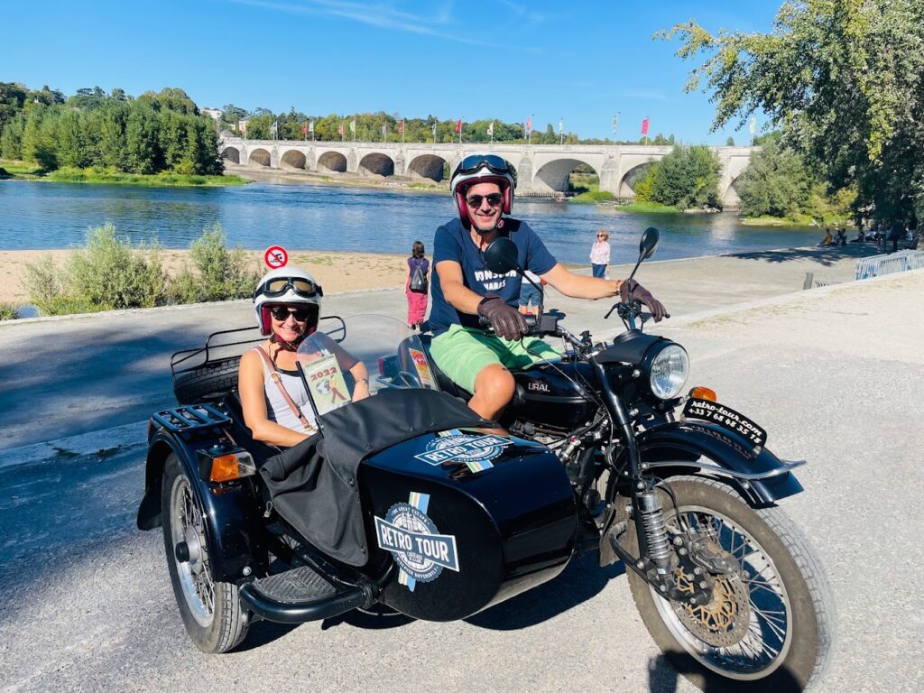 A smiling couple on a black sidecar motorcycle by the river with a historic stone bridge in the background, showcasing a unique and scenic experience on a motorcycle tour in France.