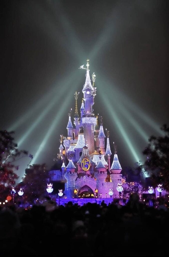 The enchanting Sleeping Beauty Castle at Disneyland Paris lit up at night with twinkling lights and a '15th anniversary' sign, bathed in dramatic beams of light, creating a magical atmosphere for a 'Disneyland Paris on a Budget' experience.