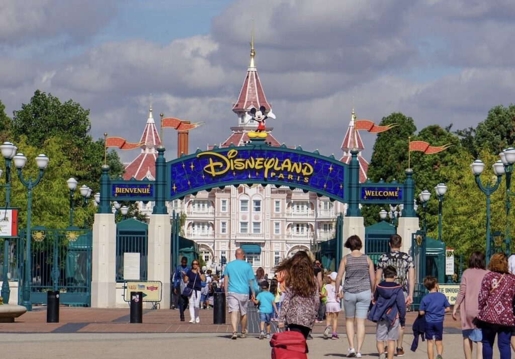 Visitors approaching the vibrant entrance of Disneyland Paris under a clear blue sky, with the iconic 'Bienvenue' and 'Welcome' signs inviting guests to the magical theme park. This image captures the essence of Disneyland paris on a budget.