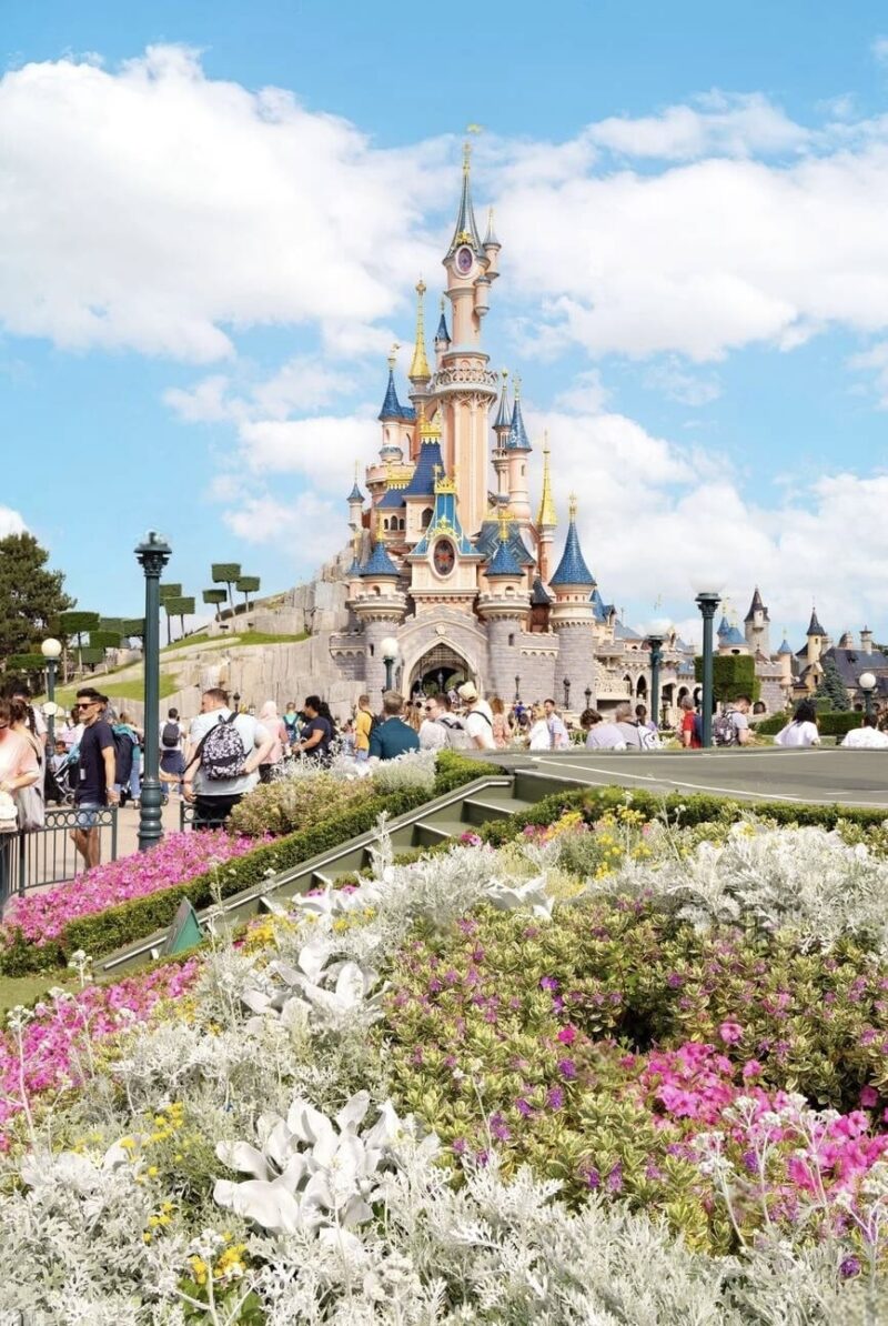 Visitors exploring around the iconic Sleeping Beauty Castle amidst vibrant floral arrangements at Disneyland Paris, capturing the essence of 'Disneyland Paris on a Budget' with its fairy tale allure.