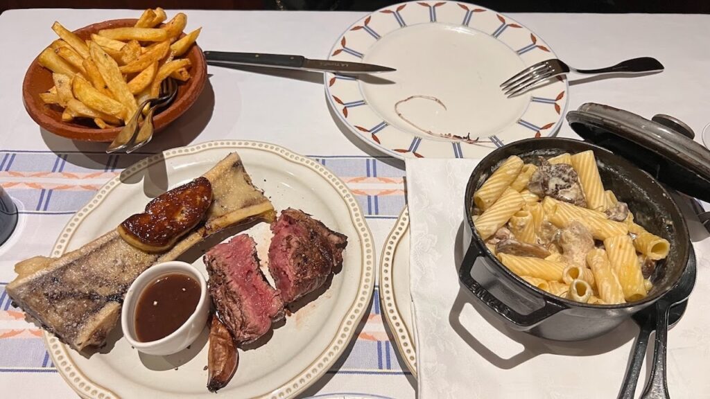 A sumptuous meal featuring Entrecôte à la Bordelaise at a local restaurant, perfect for those spending one day in Bordeaux. The table is set with a juicy steak paired with bone marrow and sauce, golden fries in a terracotta bowl, and a hearty pot of creamy pasta.