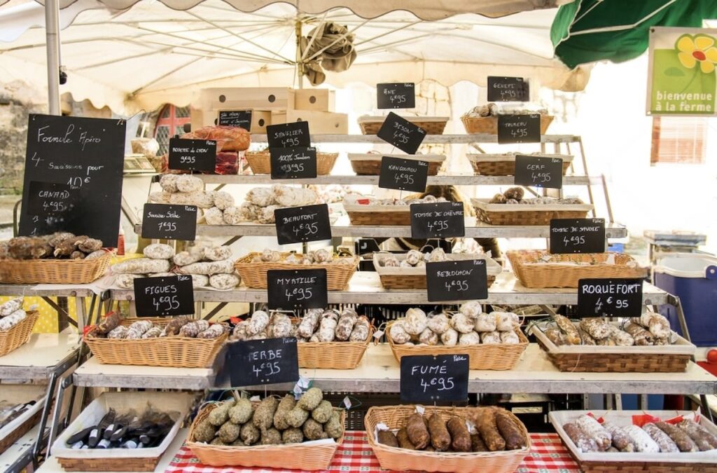 Explore the culinary delights on 'food tours in Nice, France' with this image of a local market stand, featuring a variety of handmade sausages, dried herbs, and traditional French cheeses, each with a price tag, under the welcoming sign 'bienvenue à la ferme'.