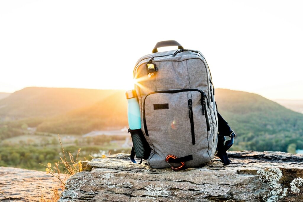 Sturdy travel backpack with a water bottle compartment, perched on a rocky outcrop against a backdrop of a picturesque sunset over hills, a crucial and versatile item on any adventurer's packing list for France.
