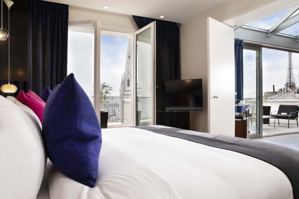 Elegant Parisian hotel room with floor-to-ceiling windows opening to a balcony with a stunning view of the Eiffel Tower, featuring a comfortable bed with crisp white linen and vibrant blue and pink pillows, appealing to searches for hotels in Paris with Eiffel Tower views.