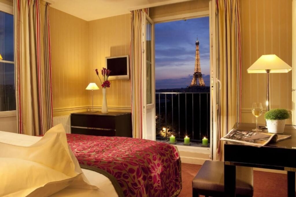 Cozy hotel room at dusk with yellow walls and striped curtains, showcasing a bed with a floral red bedspread and a window view of the illuminated Eiffel Tower, capturing the essence of hotels in Paris with Eiffel Tower views.