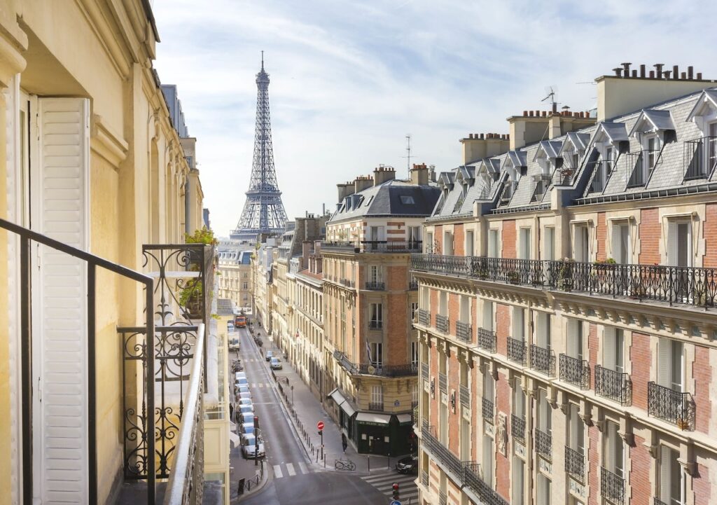 View from a Paris hotel balcony showcasing the iconic Eiffel Tower in the distance, with classic Parisian architecture lining the street below, under a clear blue sky—ideal for those seeking hotels in Paris with Eiffel Tower views.