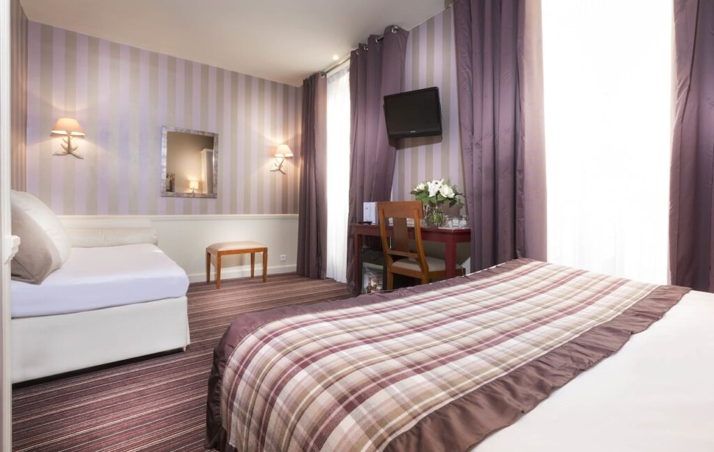 Charming hotel room in Paris decorated with lilac and white striped walls, featuring comfortable bedding with a plaid throw and a small work area, complete with fresh flowers, conveying a cozy atmosphere for visitors seeking hotels in Paris with Eiffel Tower views.