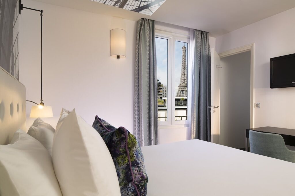 Minimalist and serene hotel room with plush white bedding and decorative pillows, wall-mounted bedside lamps, and a window that frames the majestic Eiffel Tower, ideal for those seeking hotels in Paris with Eiffel Tower views.