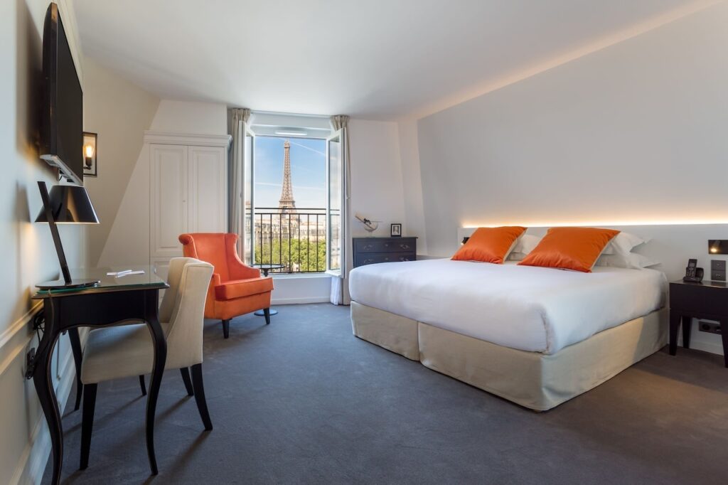 Sophisticated hotel room with a bright and airy feel, showcasing a plush bed with white linens and orange accents, an inviting orange armchair, and a large window offering a panoramic view of the Eiffel Tower, perfect for those looking for hotels in Paris with Eiffel Tower views.