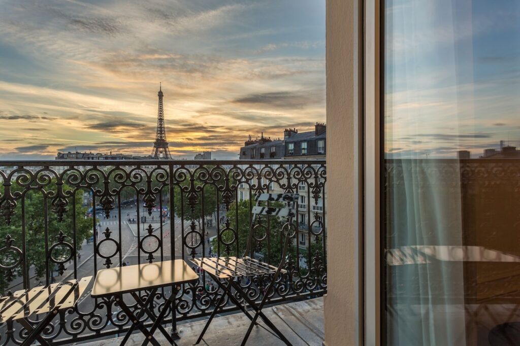 Sunset view from a wrought-iron balcony of a Paris hotel, with a bistro table and chairs overlooking the Eiffel Tower and the city's rooftops, creating a romantic setting that captures the essence of hotels in Paris with Eiffel Tower views.