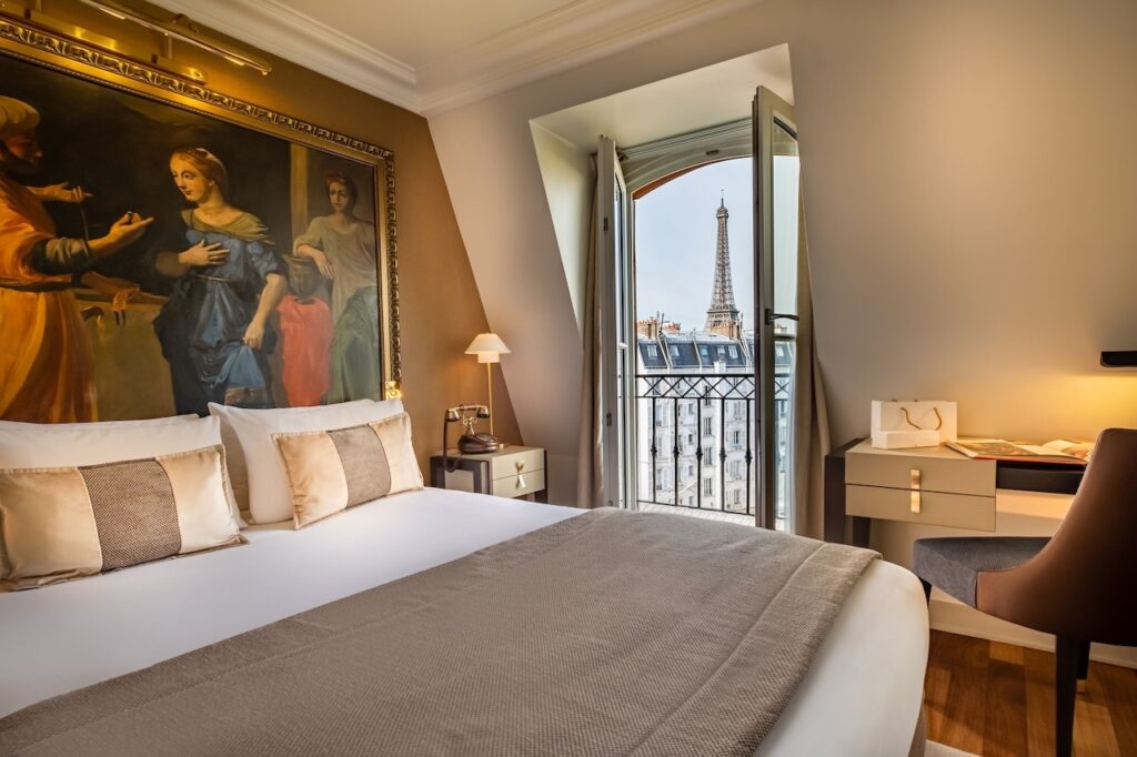 Elegant hotel room adorned with classic artwork, featuring a large comfortable bed with gold and white pillows, a sophisticated work desk, and an open French window revealing a picturesque view of the Eiffel Tower, embodying the sought-after hotels in Paris with Eiffel Tower views.