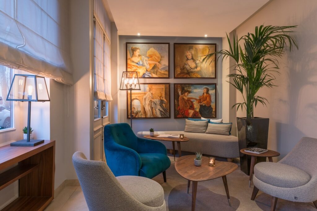 A cozy hotel lounge with an art gallery feel, featuring a plush teal armchair, soft lighting, and a collection of classic paintings, complemented by a serene sitting area with a sofa and wooden tables, showcasing the art-inspired ambiance of hotels in Paris with views of the Eiffel Tower.