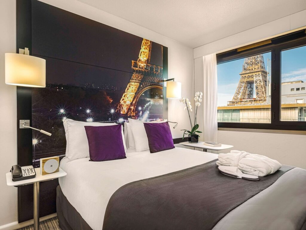 Hotel room with a stunning wall-sized photo of the Eiffel Tower at night above the bed, and a real view of the tower through the window, complete with a comfortable bed with white linens and purple accent pillows, capturing the essence of hotels in Paris with Eiffel Tower views.