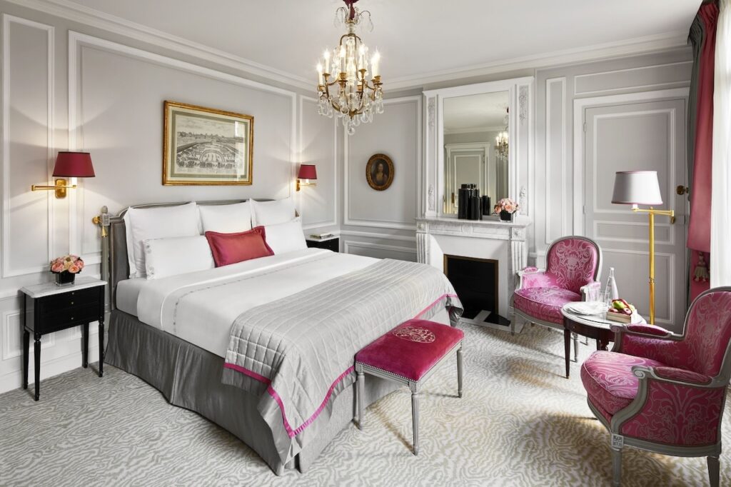 Luxuriously appointed Parisian hotel room with a plush bed, elegant white and grey bedding with pink accents, ornate chandeliers, and vintage-inspired furniture, reflecting the opulent style of hotels in Paris near the Eiffel Tower.