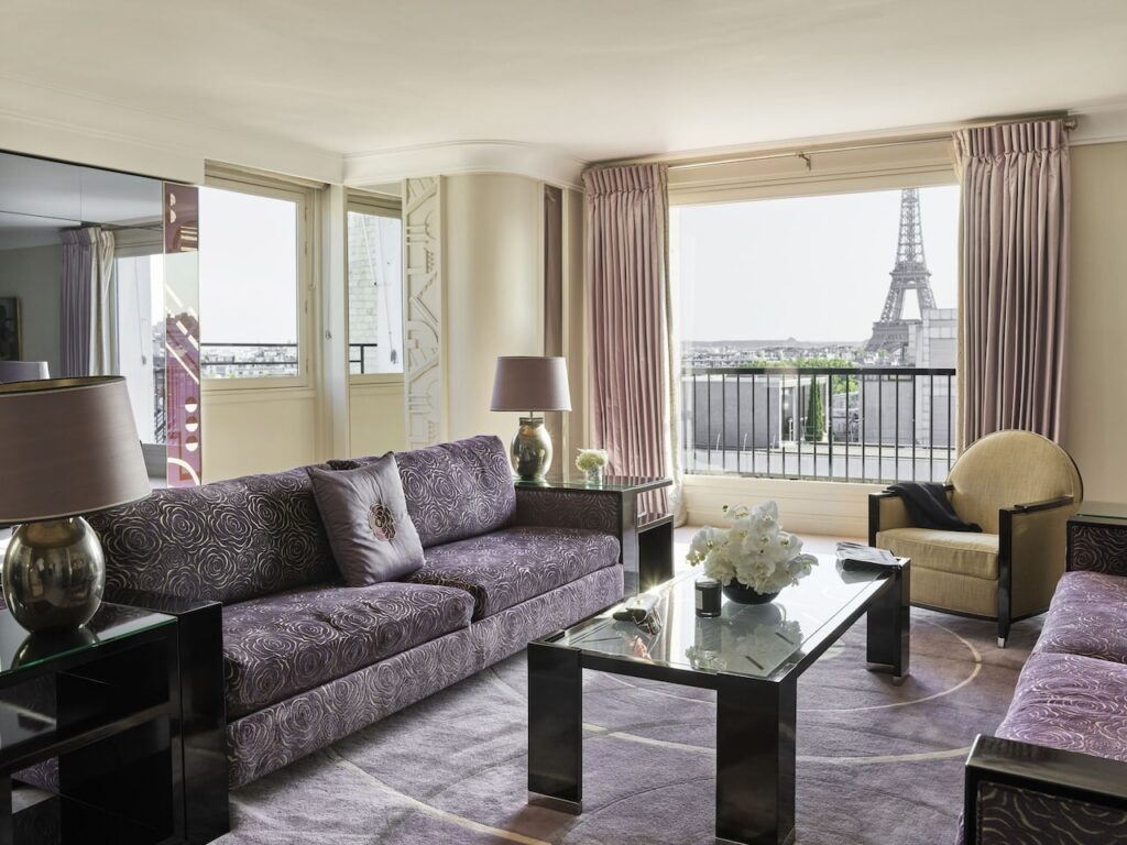 Spacious and elegantly furnished hotel living room with a large window offering a majestic view of the Eiffel Tower, adorned with a luxurious purple sofa, chic decor, and fresh flowers, encapsulating the exclusive experience offered by hotels in Paris with Eiffel Tower views.