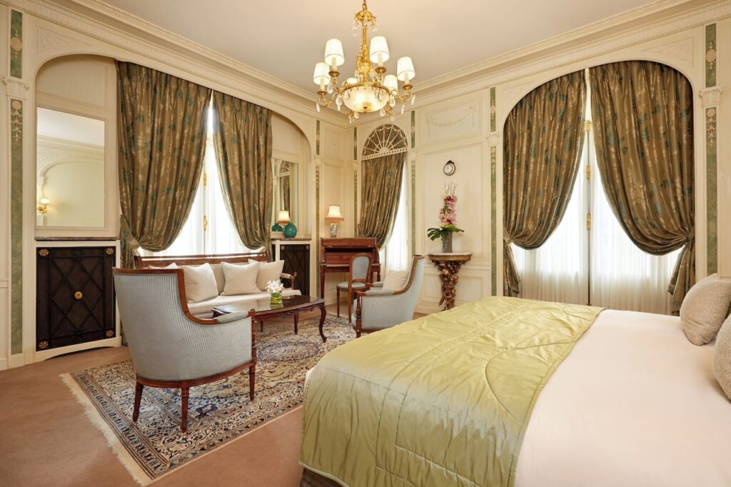 Classic French hotel room with a regal ambiance, featuring a large bed with a golden comforter, antique furnishings, ornate chandelier, and elegant drapery, offering a glimpse into the timeless luxury of hotels in Paris near the Eiffel Tower.