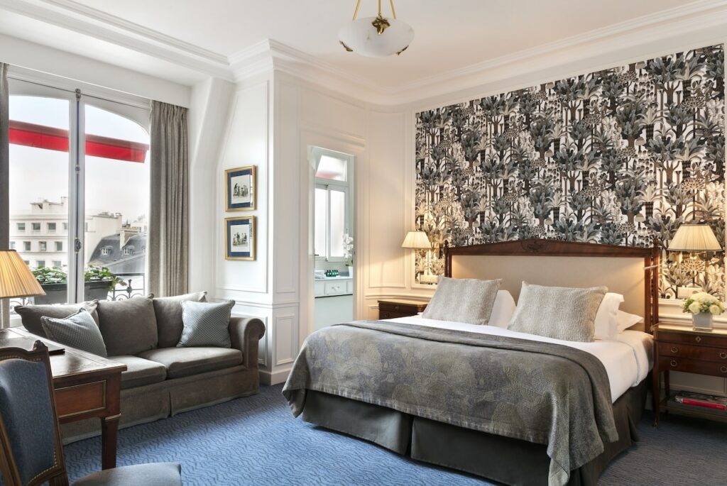 Elegant hotel room with a luxurious bed, richly patterned accent wall, and a cozy seating area by the window, bathed in natural light, offering a sophisticated retreat within a hotel in Paris, and suggesting proximity to the iconic Eiffel Tower views.