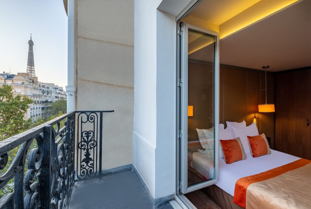 Hotel room view from a balcony showing the Eiffel Tower in the distance, featuring a bed with crisp white linens and warm orange accents, offering guests a glimpse of Parisian charm at hotels near the Eiffel Tower.