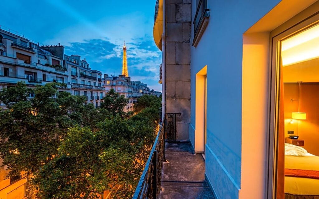 Twilight view from a hotel balcony with a narrow glimpse of the Eiffel Tower sparkling in the distance, the room's warm interior lights complementing the cool evening sky, a scene that speaks to the romantic allure of hotels in Paris with views of the Eiffel Tower.