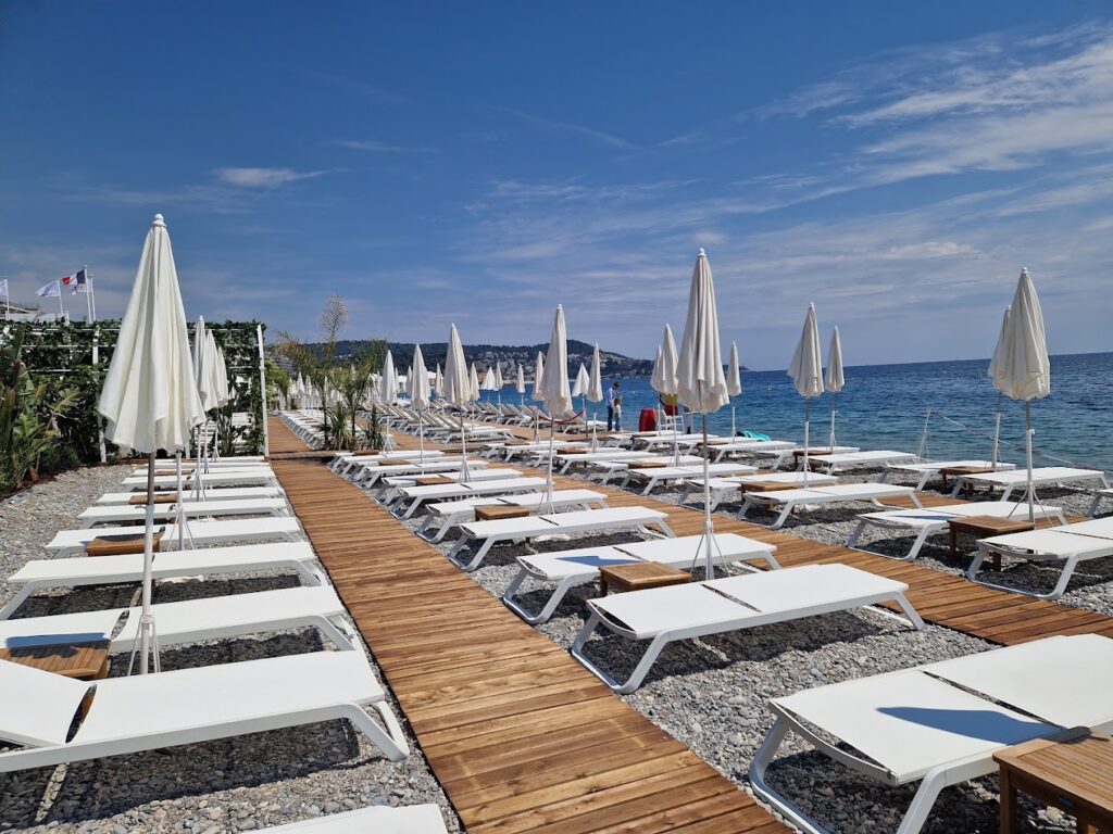 An inviting scene at one of the best Beach Clubs in Nice, with a symmetrical array of white loungers and umbrellas poised on a pebble beach. A wooden pathway leads the way towards the calm blue sea, flanked by lush greenery, under a cloudless sky—offering a tranquil Riviera experience.