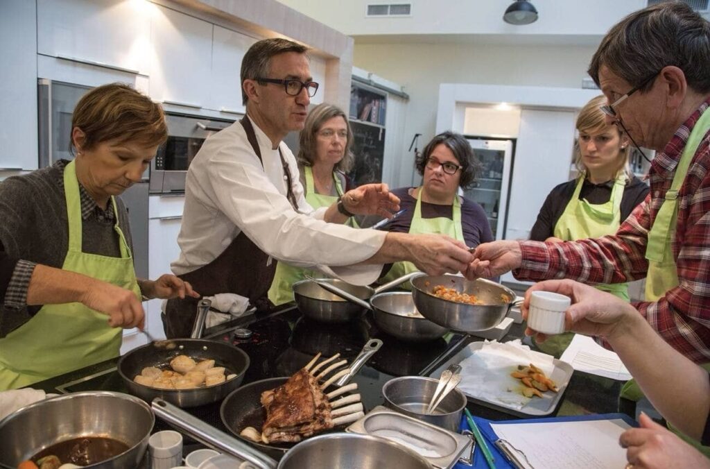 An interactive cooking class scene with participants wearing green aprons learning from a chef, surrounded by sizzling pans and ingredients, capturing the hands-on culinary experiences offered in Nice, France