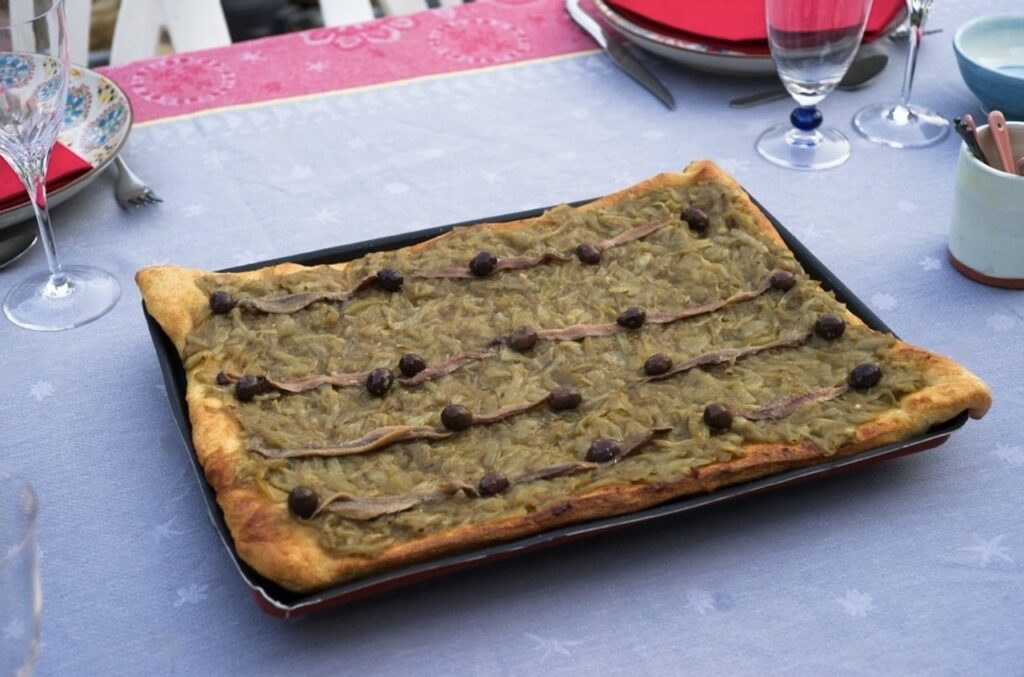 Indulge in a slice of Pissaladière, a classic dish featured on 'food tours in Nice, France', this savory caramelized onion tart is adorned with anchovies and olives, presented on a festive tablecloth, epitomizing the region's traditional cuisine.
