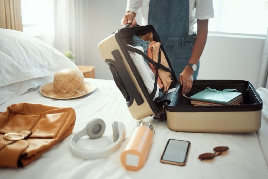 Essentials for a trip to France laid out on a bed, with a person packing clothes into a suitcase, next to a stylish straw hat, a leather purse, headphones, a water bottle, books, and sunglasses, illustrating an ideal packing list for France.